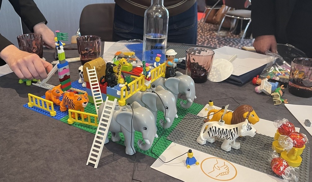 Close-up of a LEGO zoo build on a workshop table, with elephants, tigers, lions, a zebra, ladders, fences and participants' hands around the shared model.