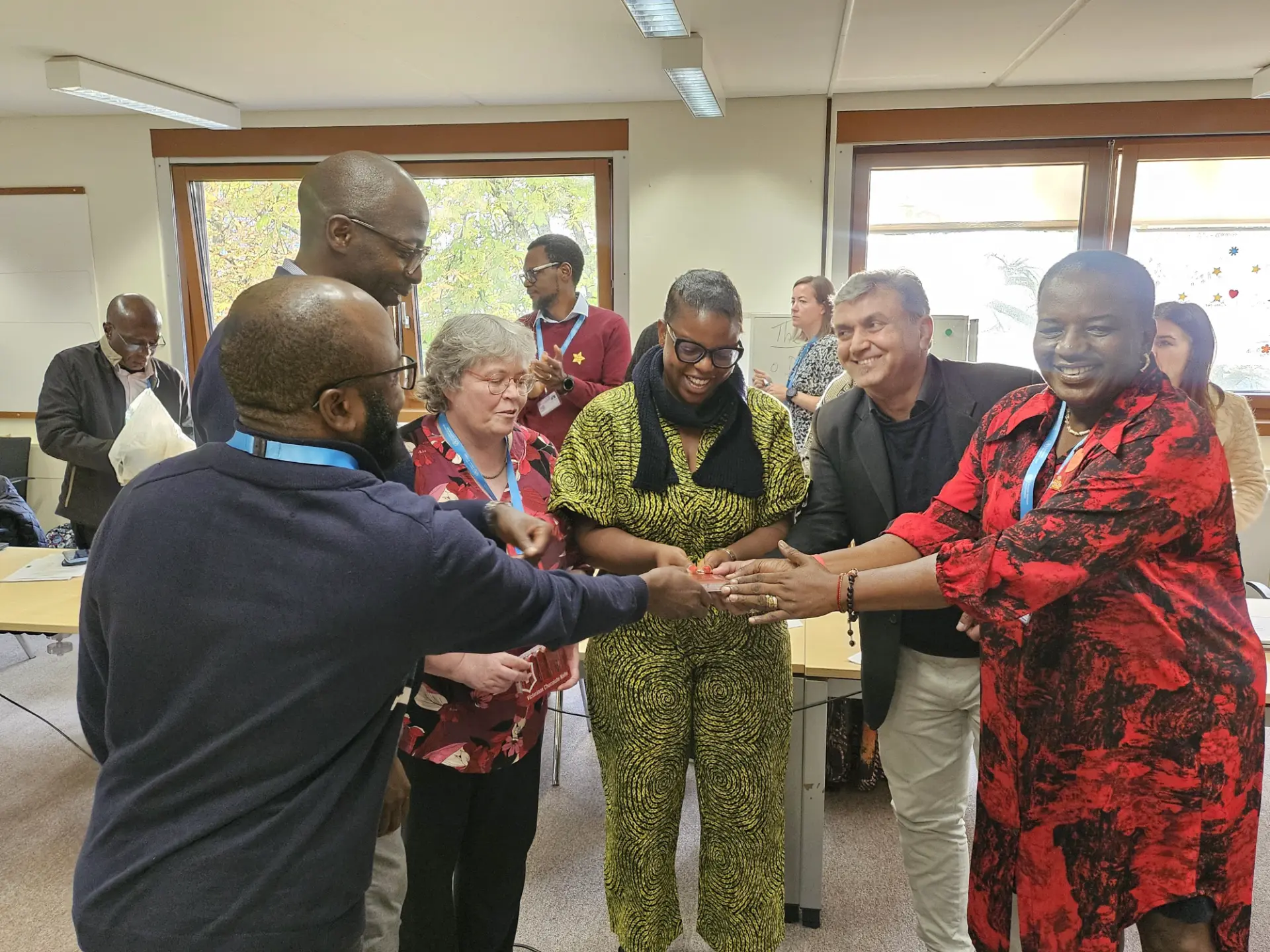A diverse group of workshop participants standing together, smiling and passing an object between them during a session.